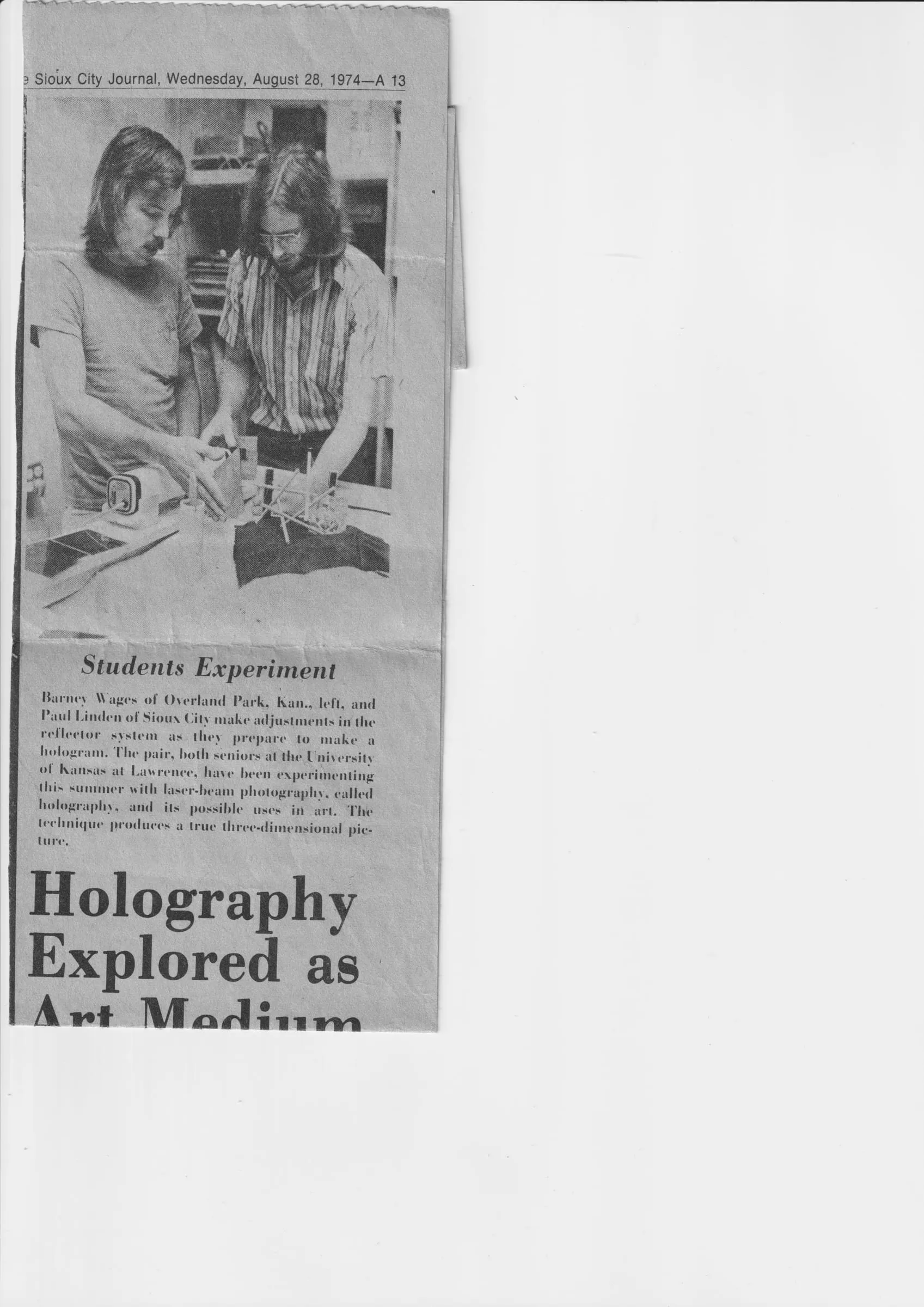 A 1974 Sioux City Journal clipping headlined "Holography Explored as Art Medium," showing two young men adjusting an optical setup to make a hologram.