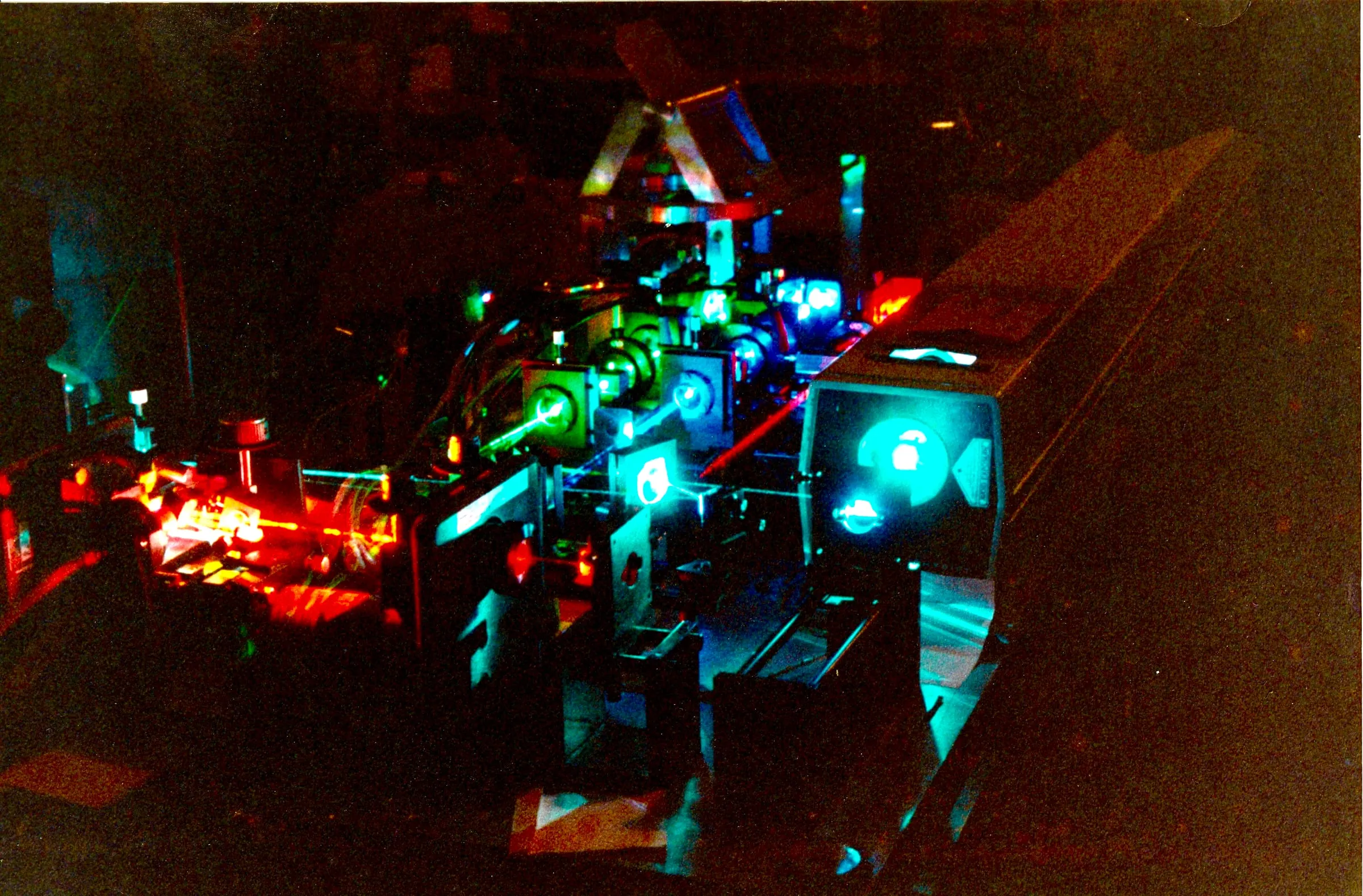 Red, green, and blue laser beams tracing through the mirrors, lenses, and modulators of a laser projection system in a darkened lab.