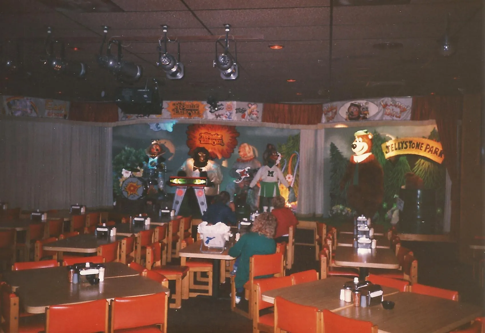 A ShowBiz Pizza Place dining room with the animatronic band performing on stage, the Jellystone Park stage at right, stage lighting overhead, and families seated at the tables.